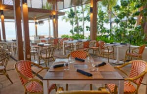 Restaurante al aire libre con sillas de mimbre y mesas, rodeado de palmeras y exuberante vegetación. Ideal para eventos y conferencias, ofrece vistas a una playa de arena y al océano bajo un cielo soleado, creando un ambiente tropical relajado.