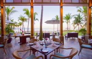 Restaurante al aire libre con mesas, sillas de mimbre y decoración tropical en un complejo turístico de México, con vistas a una playa con palmeras, sombrillas y el océano; perfecto para reuniones o eventos de conferencias bajo un cielo despejado.