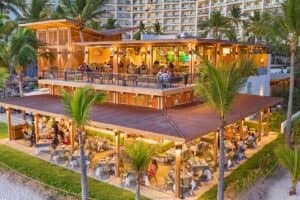Un animado restaurante frente al mar con terraza al aire libre en dos niveles, rodeado de palmeras. Ideal para reuniones o eventos, los comensales disfrutan de sus comidas bajo una iluminación cálida, con un gran hotel visible al fondo.