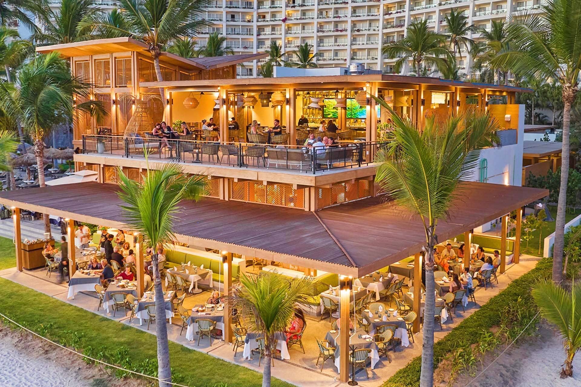 A vibrant beachfront restaurant with open-air seating on two levels, surrounded by palm trees. Perfect for meetings or conference events, diners enjoy meals under warm lighting, with a large hotel building visible in the background.