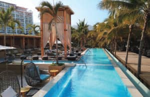 En este moderno hotel resort mexicano le espera una lujosa piscina al aire libre rodeada de palmeras, tumbonas y camas con dosel, perfecta para relajarse entre eventos o reuniones bajo un cielo despejado.
