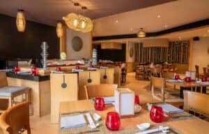 El interior de un restaurante moderno en un complejo turístico de México, con mesas y sillas de madera, vasos de cristal rojo, faroles de paja, detalles de bambú y una barra de sushi al fondo: un entorno acogedor, perfecto para reuniones o eventos de conferencias.