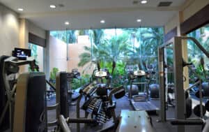 Un gimnasio moderno en los complejos turísticos de México con cintas de correr, máquinas elípticas y soportes para pesas, con grandes ventanales de cristal con vistas a exuberantes palmeras, perfecto para los huéspedes que asisten a conferencias o reuniones.
