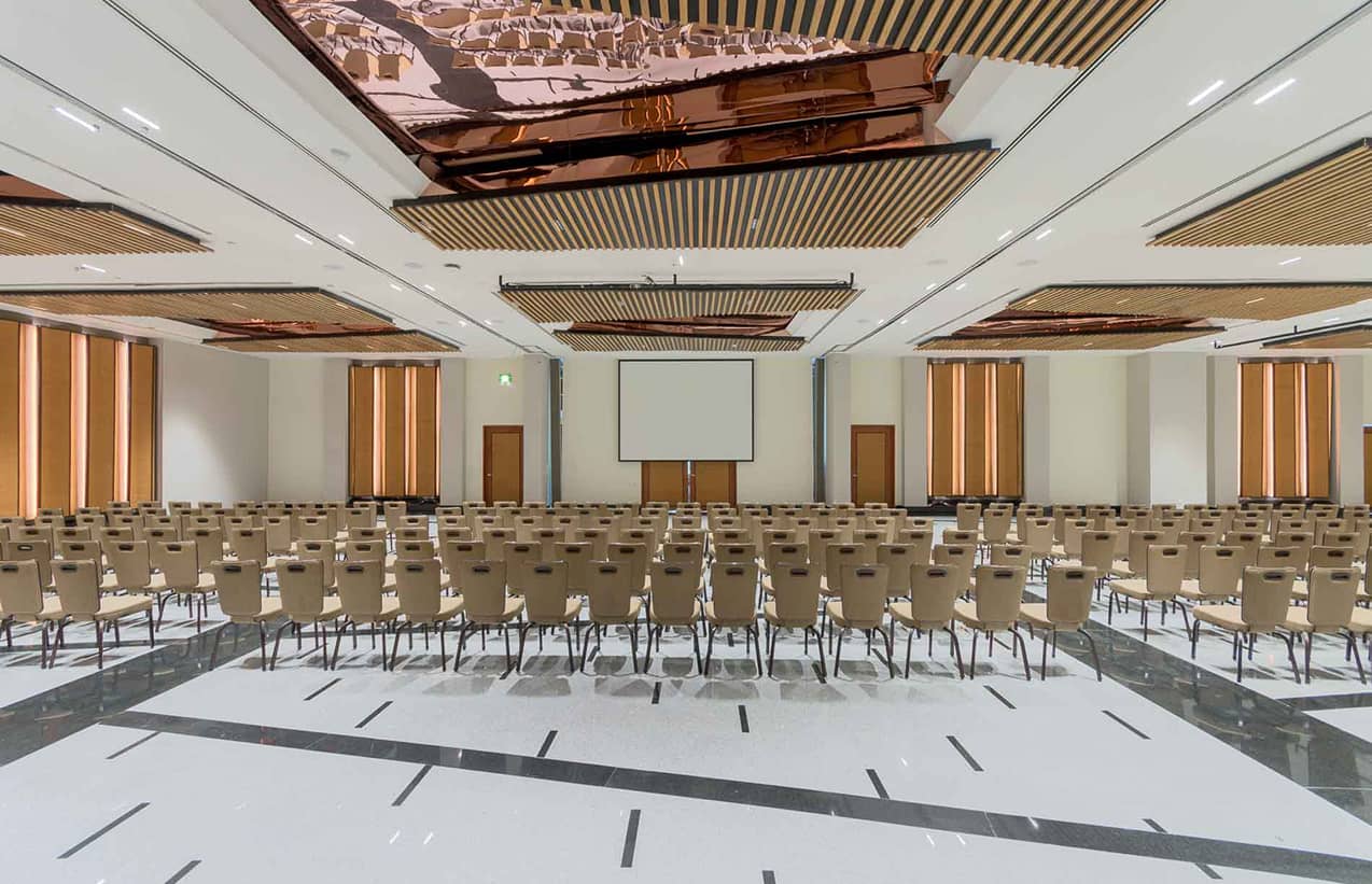 A large, modern conference room in one of Mexico's top hotels, with rows of beige chairs facing a projection screen, polished floors, and decorative ceiling panels. Ideal for meetings or conference events, the space is well-lit with natural and artificial light.