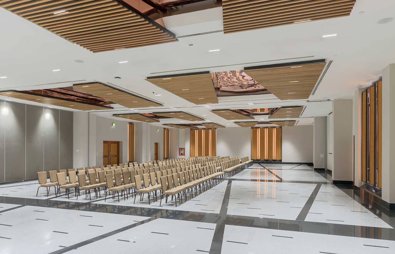 A large, modern conference room for meetings and conference events, with rows of beige chairs facing forward, wooden ceiling panels, tall windows, and a glossy white floor with black accents. The space is empty and well-lit.