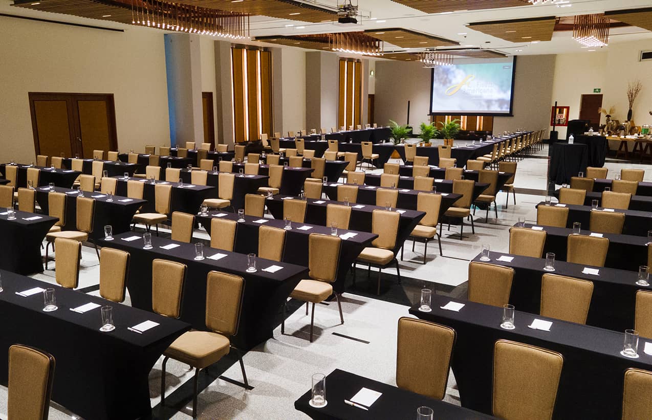 A large conference room in a Mexico hotel is set up classroom-style with rows of tables and chairs, each with a glass and notepad. A projector screen displays a presentation, perfect for meetings or conference events at top resorts.