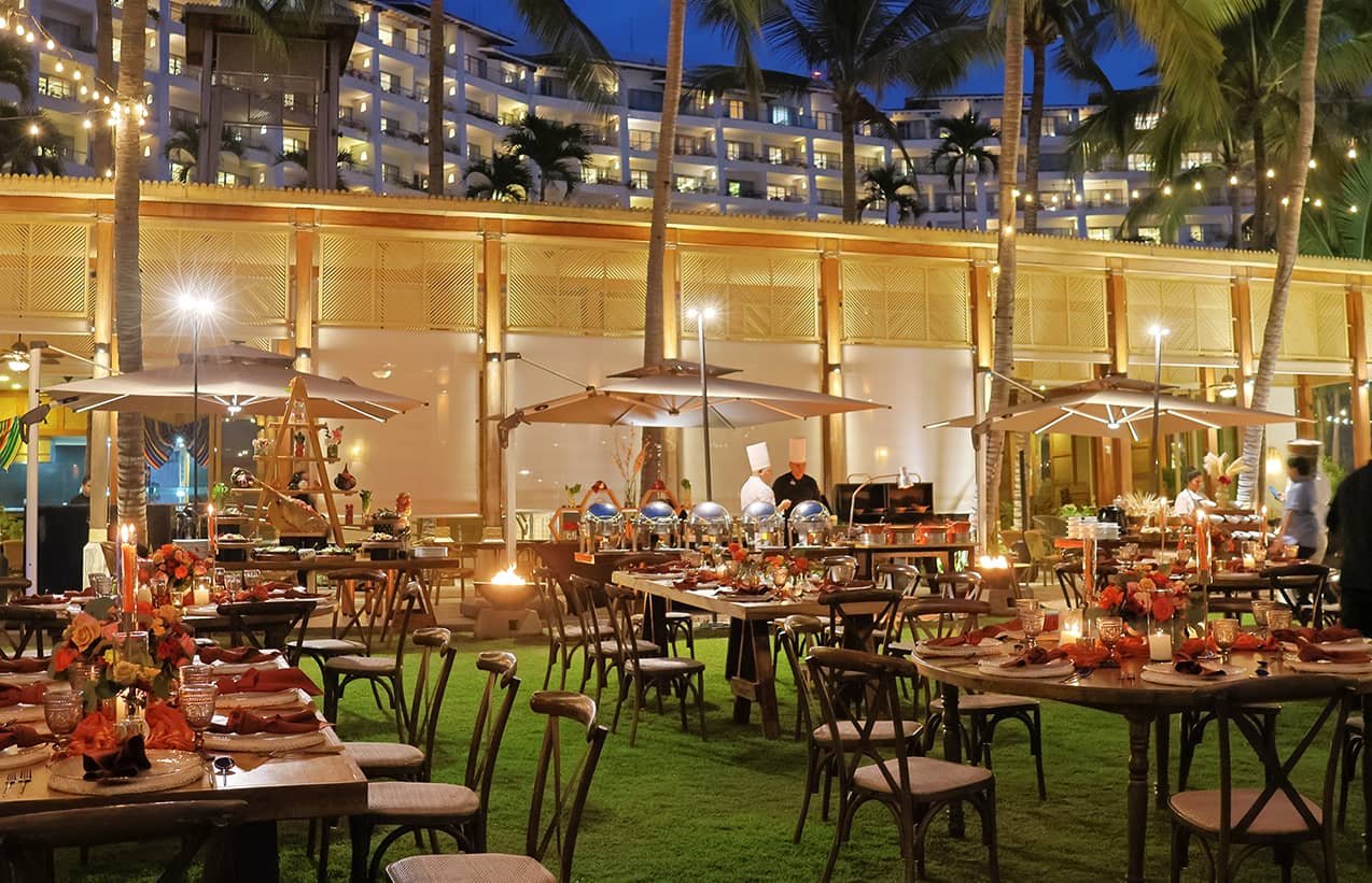 Outdoor evening conference event on a lawn with decorated tables, string lights, and buffet stations. Chefs prepare food as guests mingle under umbrellas. A lit Mexico resort hotel, balconies, and palm trees complete the scene.