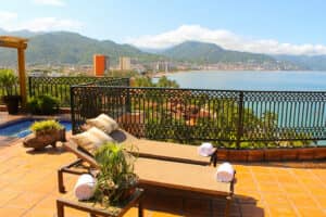 Dos tumbonas con toallas enrolladas se encuentran en una soleada terraza de un resort en México, con vistas a una ciudad costera, montañas y el océano. La terraza, cercada, cuenta con plantas en macetas y una pequeña piscina, ideal para relajarse entre eventos o reuniones.