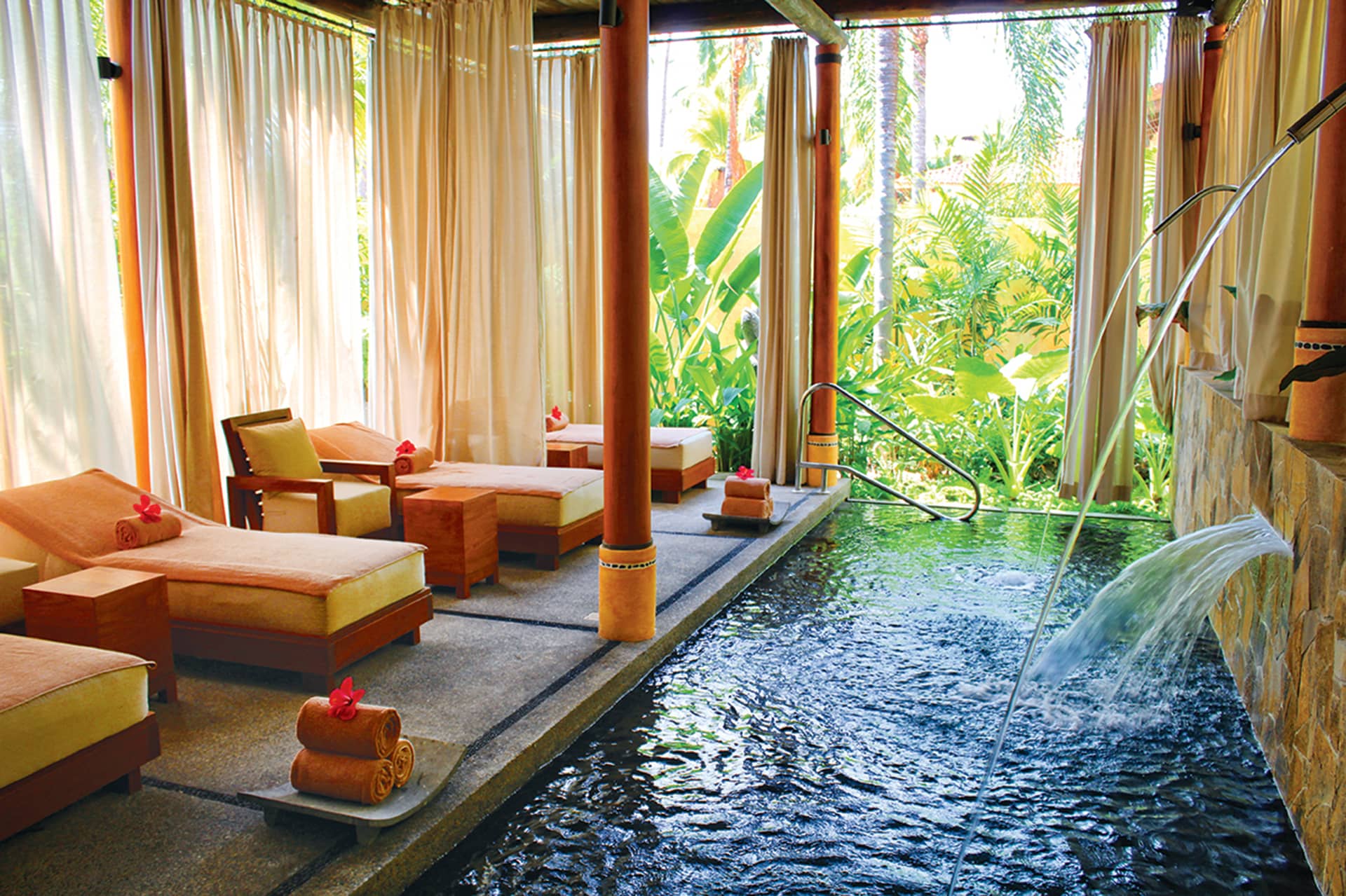 A serene spa room with cushioned lounge chairs, rolled towels, and a narrow pool awaits at one of Mexico’s top resorts. Lush greenery and palm trees outside the windows create a relaxing escape for guests between meetings or conference events.