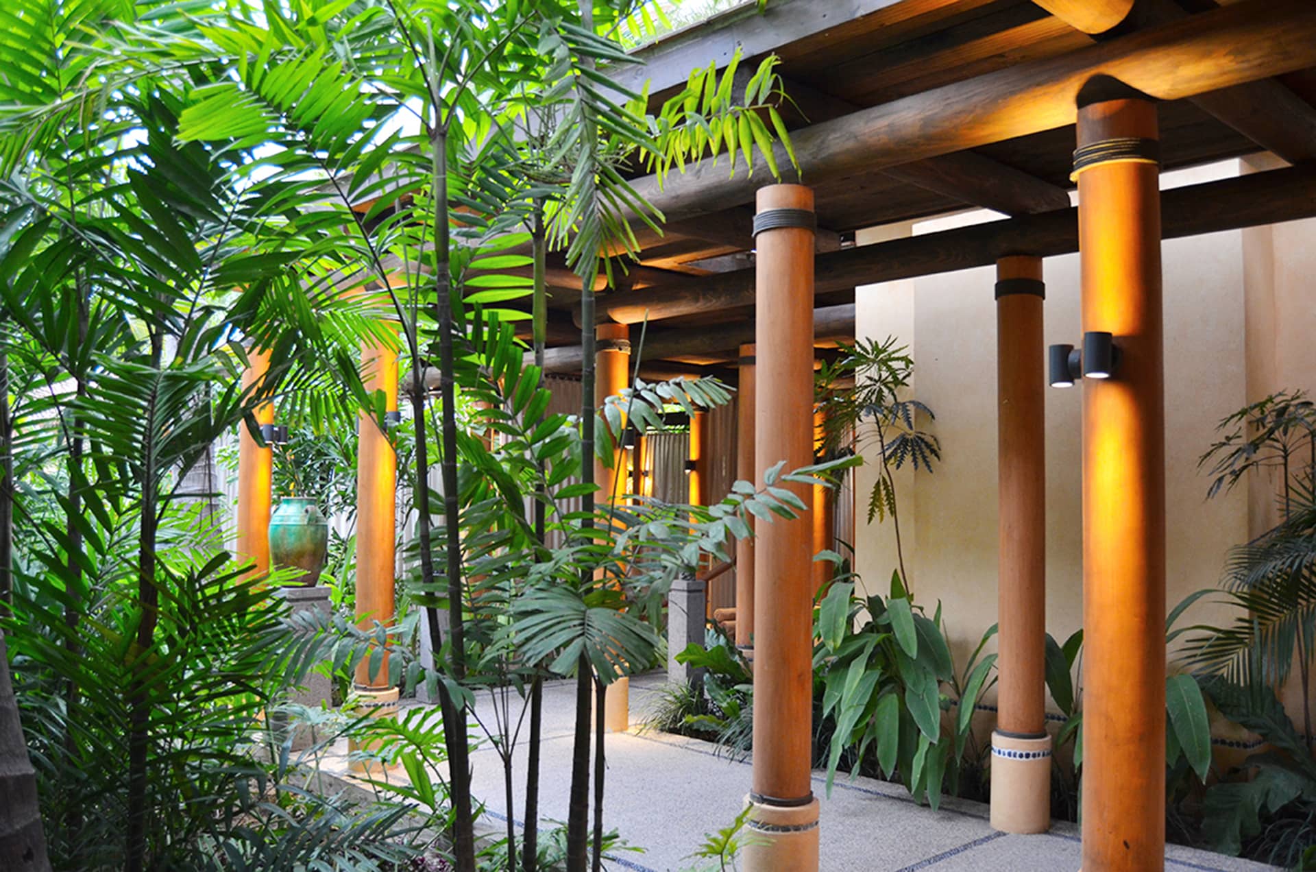 A walkway with tall wooden columns runs alongside a building at one of Mexico's premier resorts, surrounded by lush green tropical plants and trees, creating a serene and inviting atmosphere perfect for meetings or conference events.
