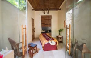 Una serena sala de spa en uno de los mejores resorts de México, con una camilla de masajes cubierta con telas marrones y doradas, vigas de madera, puertas de cristal, flores frescas y una cálida luz natural: el refugio perfecto después de eventos o reuniones de negocios.
