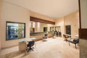 Salón luminoso y moderno con tres estaciones de trabajo, sillas negras, espejos y suelos de mármol claro: ideal para conferencias o reuniones en complejos turísticos. Estanterías con productos y un gran ventanal con vistas al exterior crean un espacio ordenado y acogedor.