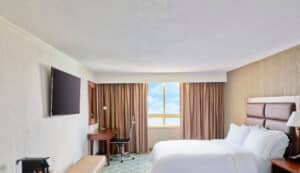 Una moderna habitación de hotel en México con una cama impecablemente hecha, televisor montado en la pared, escritorio con silla y lámpara, un banco y una gran ventana con cortinas marrones que dejan entrar la luz natural; perfecta para relajarse después de reuniones o eventos de conferencias.