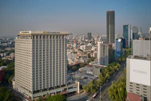 Vista aérea de un paisaje urbano con edificios altos, entre los que destaca el emblemático hotel FIESTA AMERICANA —ideal para conferencias y reuniones—, rodeado de árboles y calles concurridas. El cielo está despejado y presenta una ligera bruma.