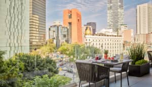La terraza en la azotea de uno de los mejores hoteles de México ofrece una mesa puesta para dos con fruta, bebidas y desayuno, con vistas a una calle concurrida y a modernos rascacielos; perfecta para relajarse después de eventos o reuniones.