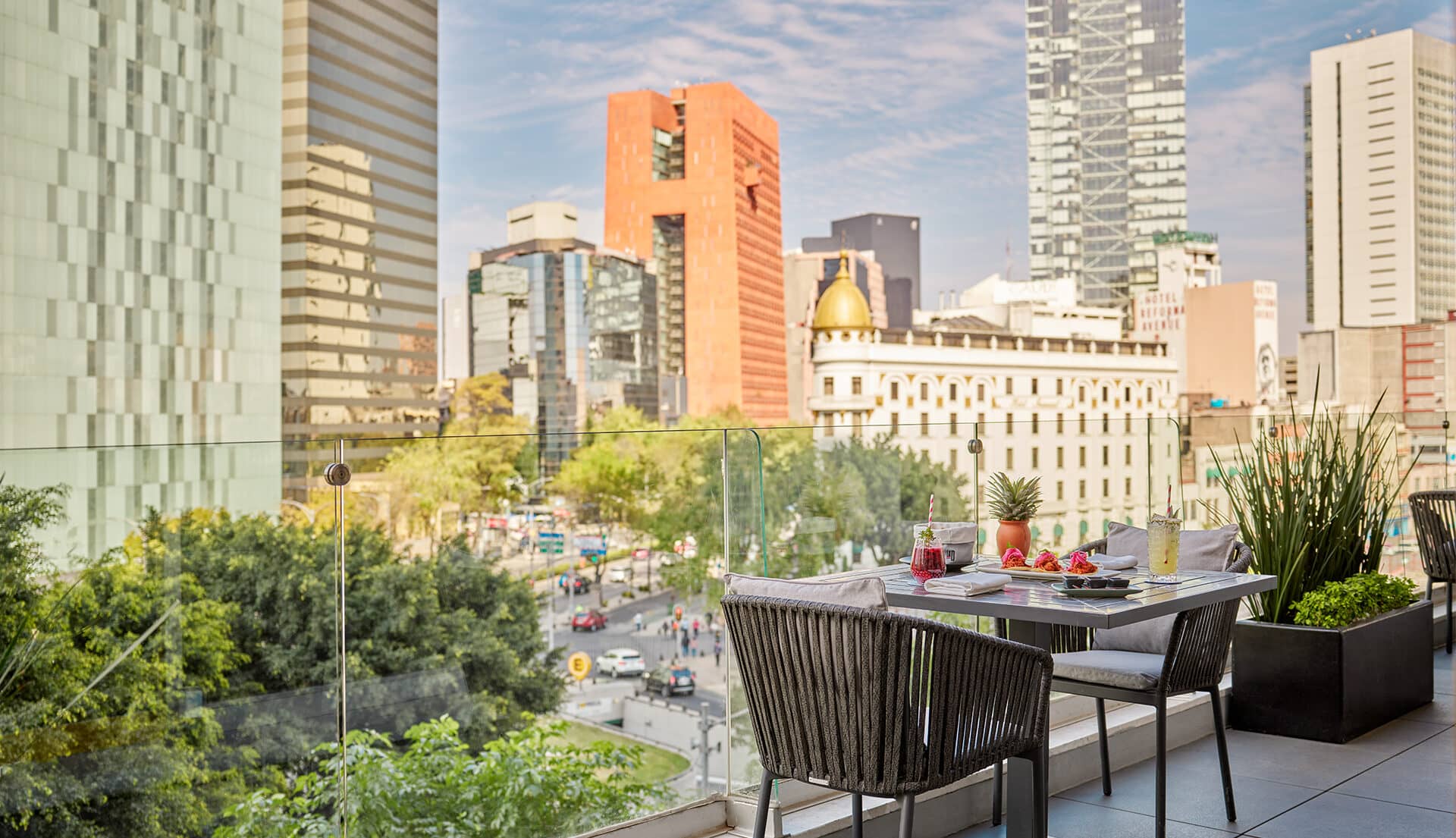 A rooftop patio at one of Mexico’s finest hotels offers a table set for two with fruit, drinks, and breakfast food, overlooking a busy street and modern skyscrapers—perfect for relaxing after conference events or meetings.
