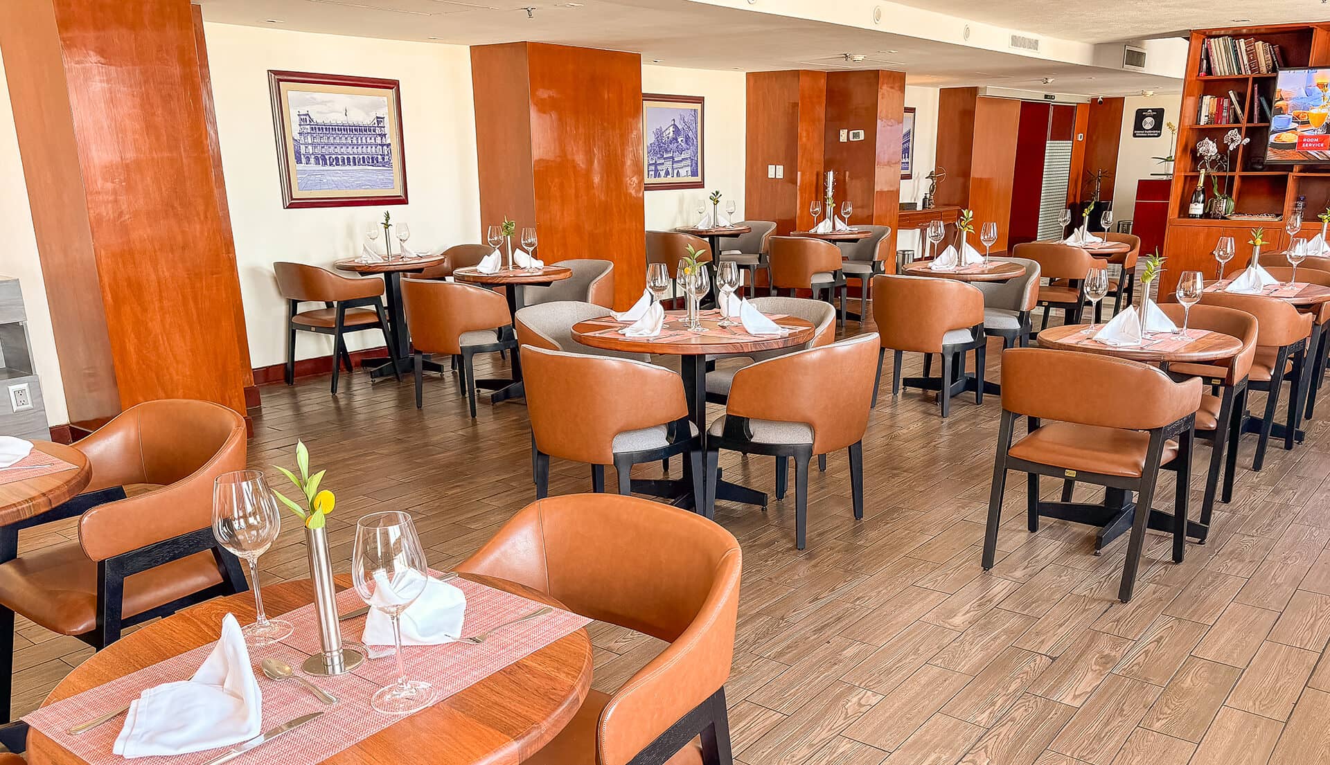 A modern restaurant dining area with round wooden tables, brown leather chairs, white napkins, wine glasses, and minimalist decor—ideal for meetings or conference events in upscale Mexico hotels and resorts. Warm wood floors and framed art accent light brown walls.