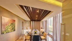 Comedor moderno con una mesa larga para doce personas, sillas acolchadas y grandes ventanales con cortinas transparentes: ideal para reuniones o conferencias. El techo contemporáneo con detalles en madera y obras de arte abstracto crea un ambiente sofisticado, similar al de un resort. La luz natural inunda el espacio.