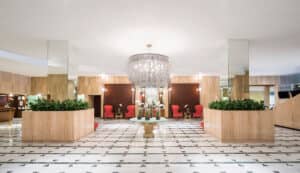 Amplio vestíbulo de hotel, ideal para reuniones, con una gran lámpara de araña de cristal, suelos de mármol con motivos geométricos, columnas con espejos, plantas verdes, sillas rojas y paredes revestidas de madera. Una mesa redonda con flores blancas se ubica en el centro, debajo de la lámpara de araña.