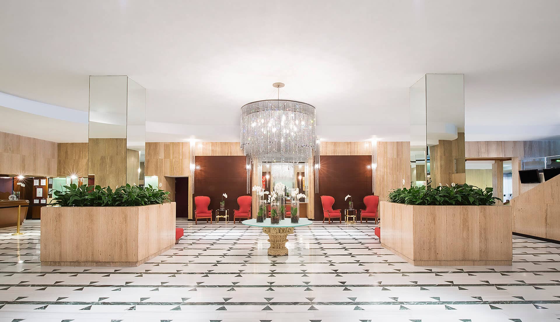 Spacious hotel lobby perfect for meetings, with a large crystal chandelier, marble floors with geometric patterns, mirrored pillars, green plants, red chairs, and wood-paneled walls. A round table with white flowers is centered beneath the chandelier.