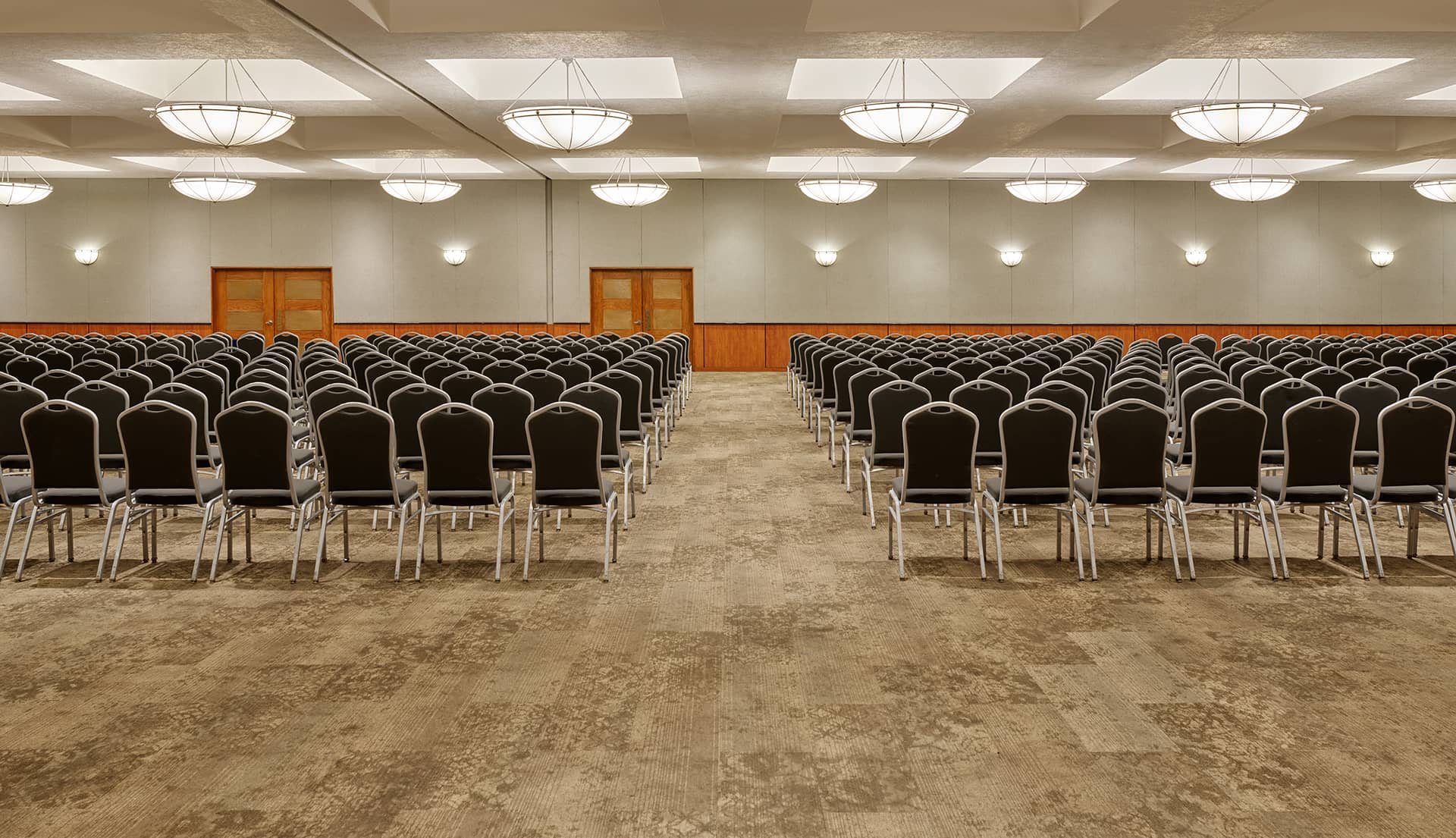 A large, empty conference room at a Mexico resort with rows of black chairs arranged theater style facing the front, beige carpeted floor, neutral walls, wooden panel accents, and several round ceiling lights—ideal for meetings or conference events.