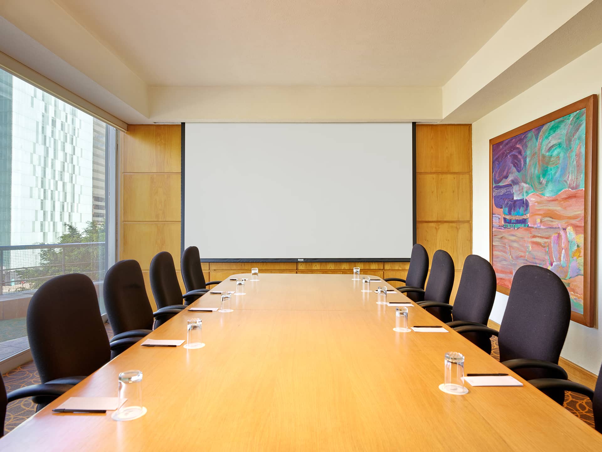 A modern conference room in Mexico resorts features a long wooden table, black chairs, notepads, pens, and glasses of water. A large blank screen and abstract art adorn the walls, with windows letting in natural light—perfect for meetings or conference events.