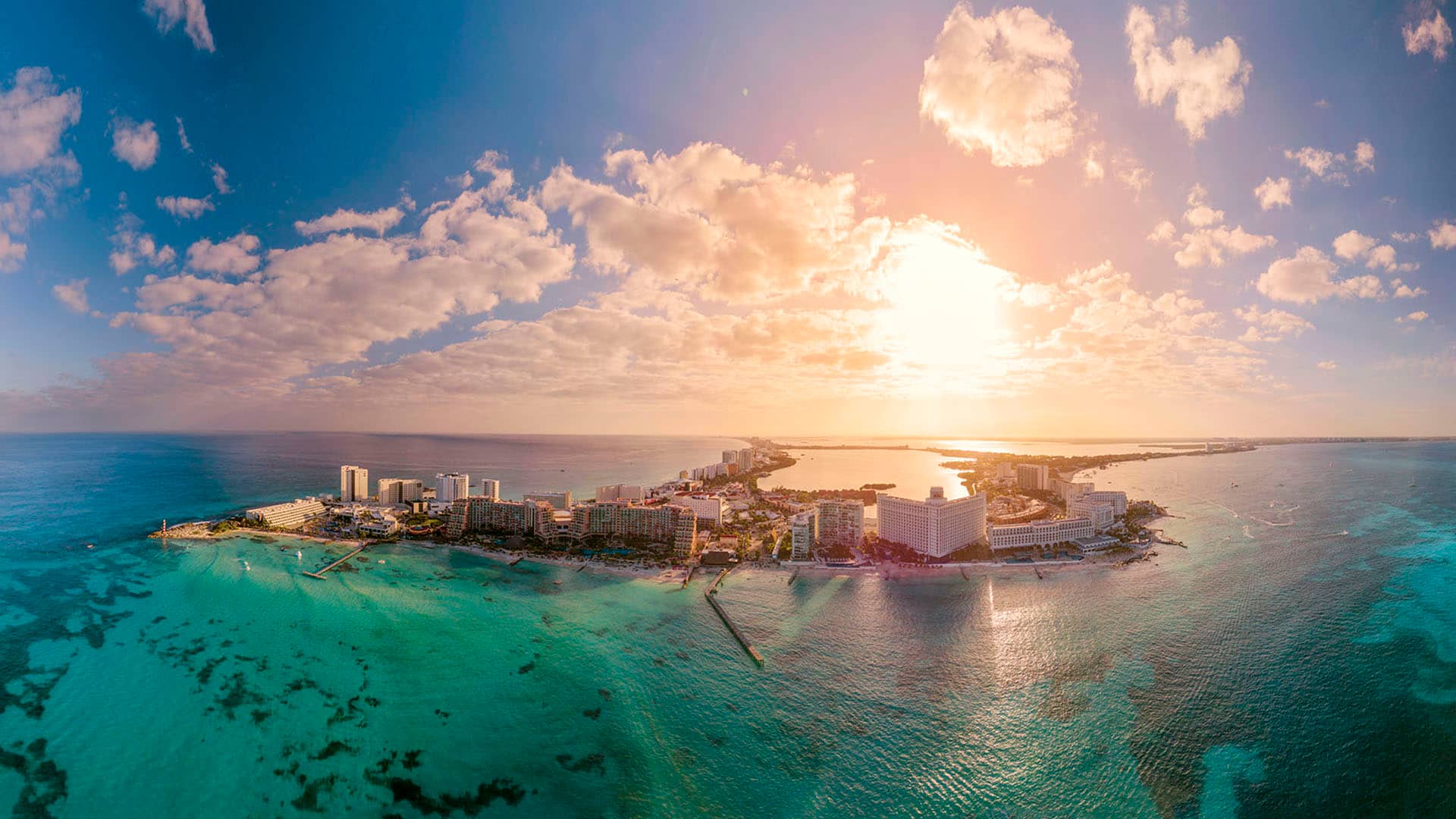 Aerial view of a coastal city in Mexico with tall hotels and conference resorts, white sandy beaches, and turquoise ocean water under a bright, partly cloudy sky at sunrise or sunset.