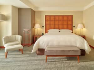 Una habitación de hotel moderna con una cama grande centrada frente a un cabecero de madera, dos mesitas de noche con lámparas y un sillón beige: perfecta para relajarse después de asistir a reuniones o eventos de conferencias en los principales hoteles y resorts de México.
