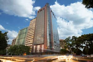 Un alto y moderno edificio de oficinas con ventanales de cristal se alza contra un cielo azul parcialmente nublado, rodeado de árboles y estelas de luz de los coches que pasan: un escenario ideal para reuniones de negocios o eventos de conferencias.