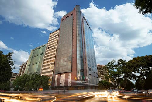A tall, modern office building with glass windows stands against a partly cloudy blue sky, surrounded by trees and light trails from passing cars—an ideal setting for business meetings or conference events.
