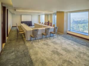 Una moderna sala de conferencias en los hoteles de México con una mesa dispuesta en forma de U, sillas alrededor de la mesa, una pantalla de proyección al frente y grandes ventanales que ofrecen vistas panorámicas de la ciudad y la vegetación, perfecta para reuniones o eventos de conferencias.