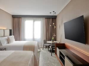 Habitación de hotel moderna con dos camas dobles, televisor de pantalla plana, mesa redonda con dos sillas (ideal para reuniones), lámparas colgantes y grandes ventanales con cortinas transparentes y opacas que dejan entrar la luz natural.