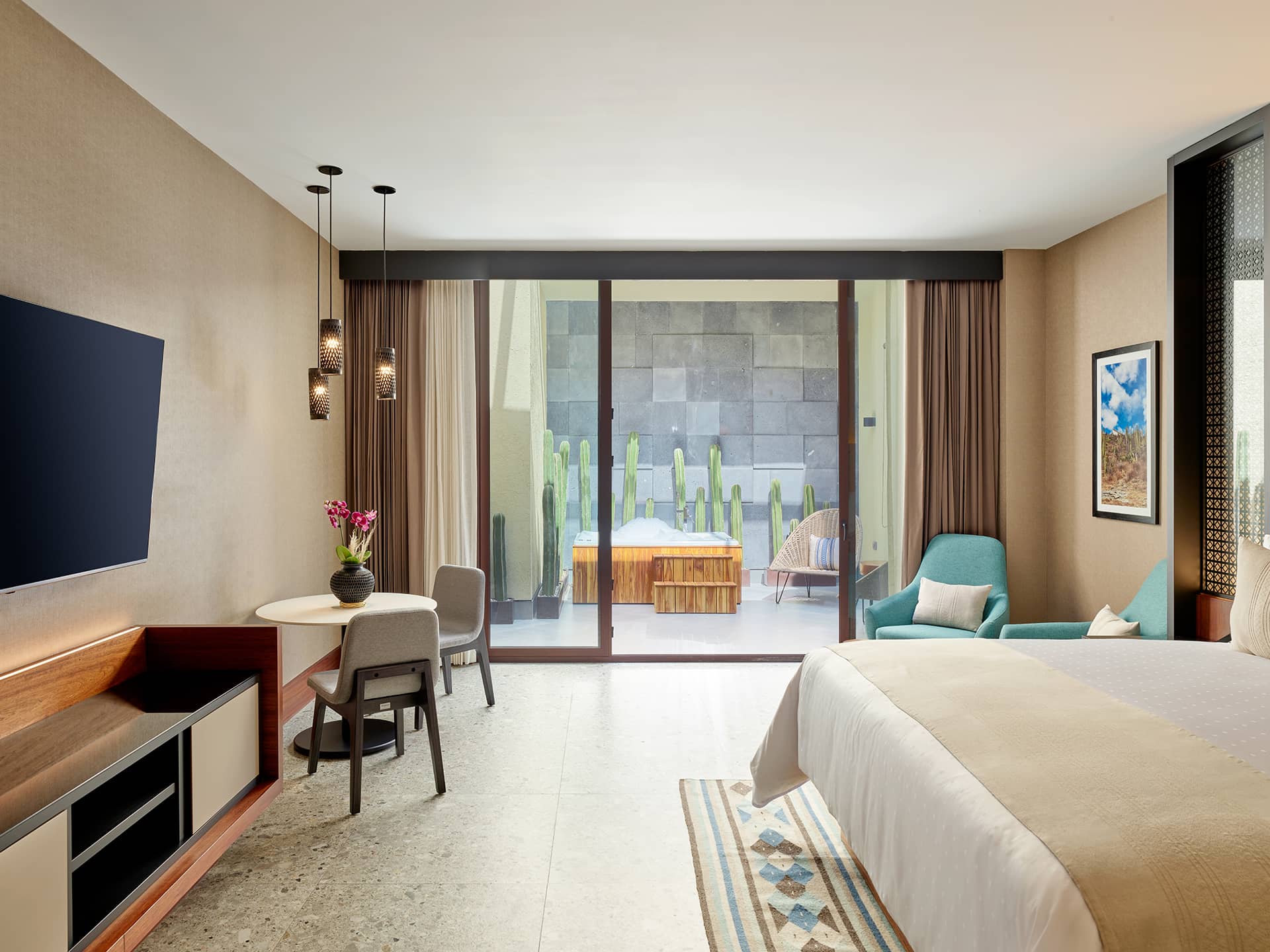 A modern hotel room at one of the top Mexico hotels, featuring a large bed, seating area, table with chairs, flat-screen TV, and sliding glass doors opening to a patio with cactus plants. Natural light fills this stylish space—ideal for meetings or relaxation.