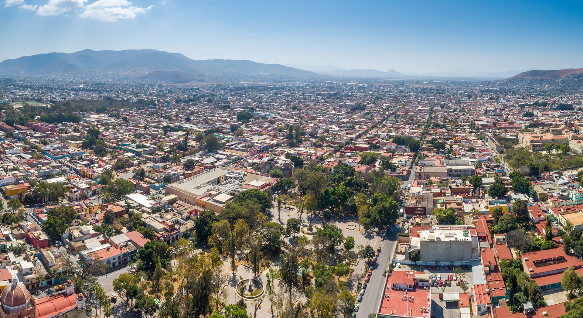 Aerial view of a sprawling city with colorful low-rise buildings, tree-lined streets, Mexico hotels near a central park area, and mountains visible in the background under a clear blue sky.