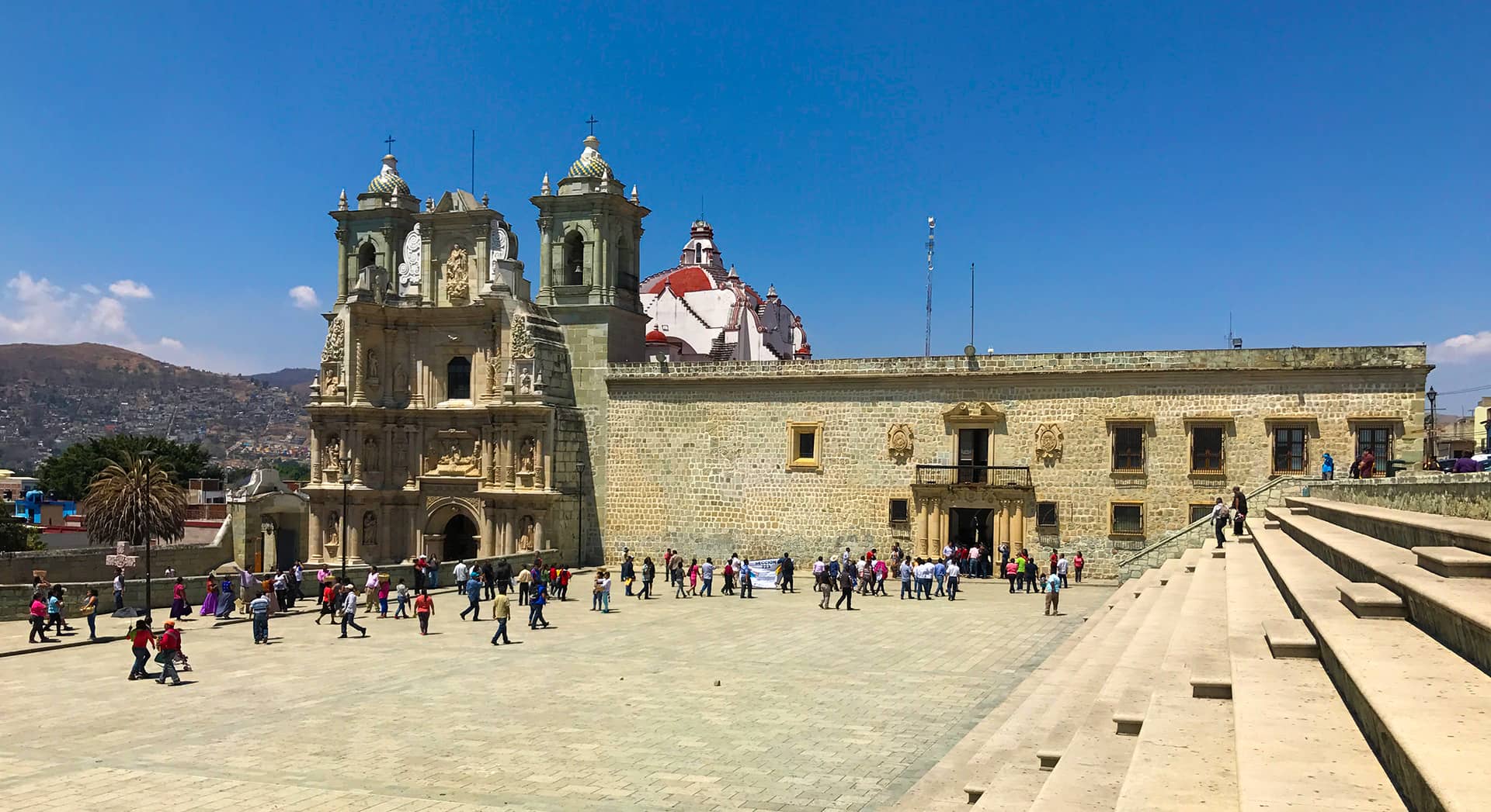 A large stone plaza with people walking and gathering in front of an ornate church with two bell towers and a dome. Stone steps are on the right, hills are visible under a bright blue sky—an inviting spot near top Mexico hotels and conference events.