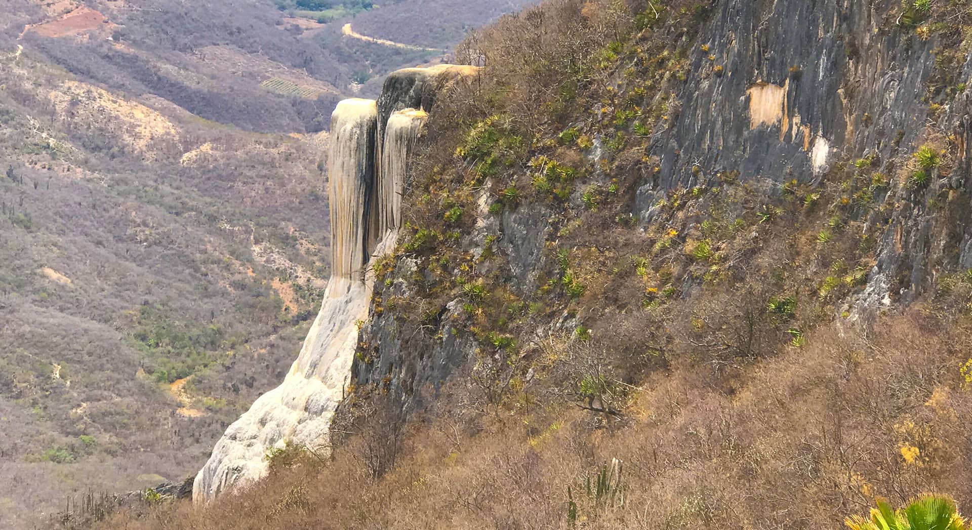A rocky cliff with mineral formations resembling a frozen waterfall rises amid dry, shrub-covered hills and mountains under a hazy sky, creating a breathtaking backdrop for conference events or retreats at resorts in Mexico.