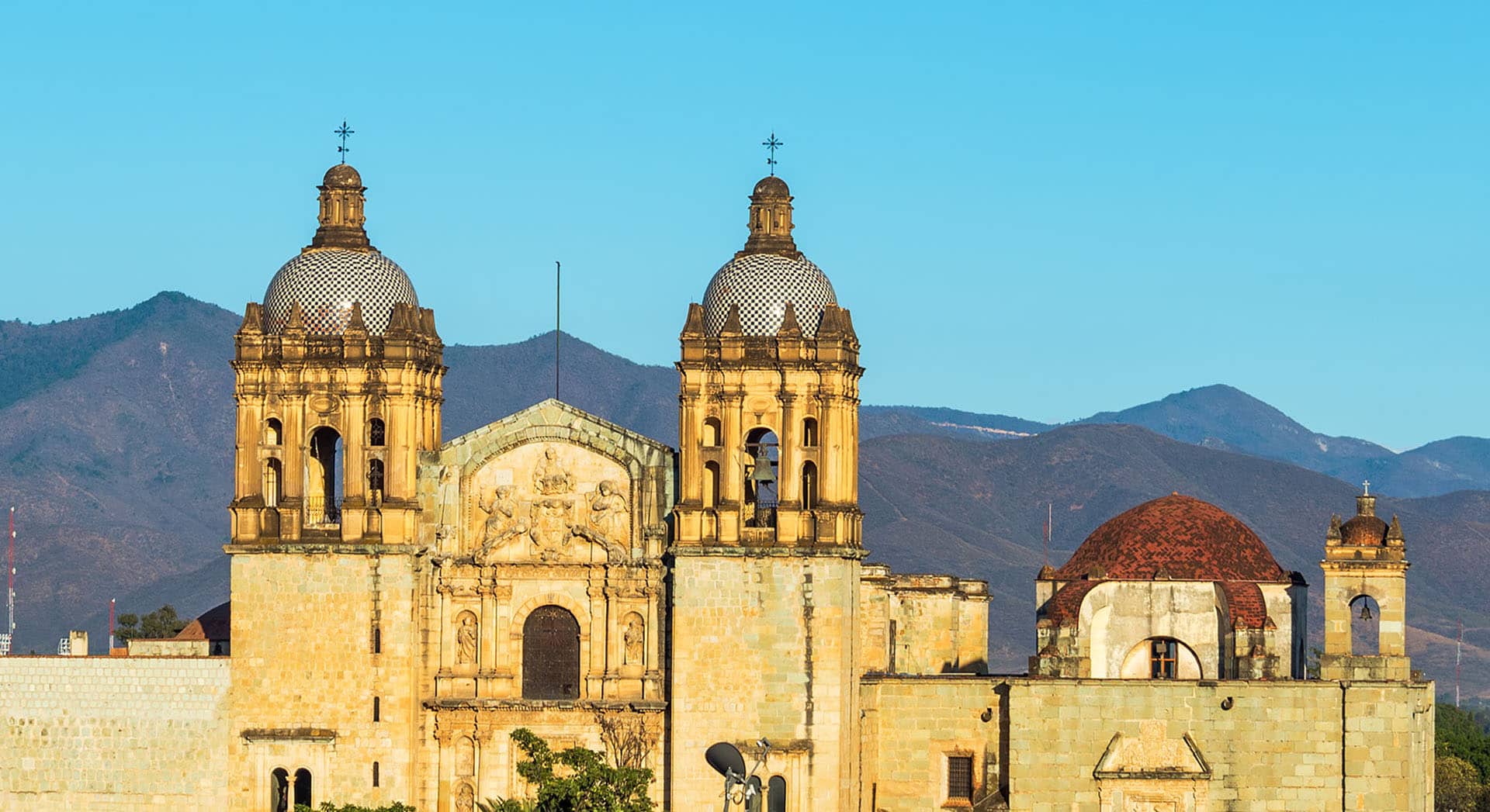 The image shows the ornate twin bell towers and dome of a historic stone church with mountains in the background, set under a clear blue sky—an iconic scene near top resorts and hotels perfect for conference events in Mexico.