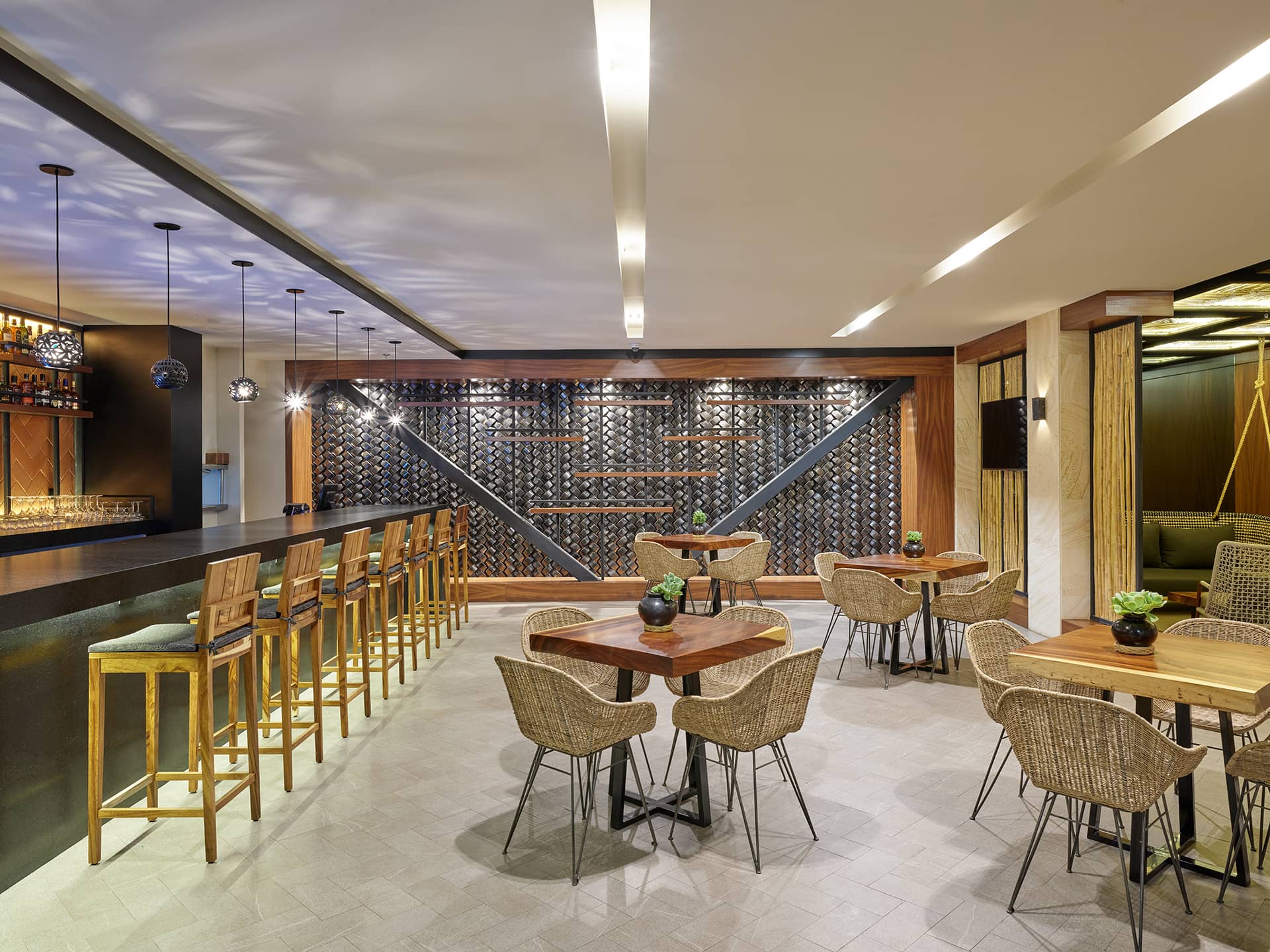 A modern bar and lounge area in Mexico resorts, with wooden tables and chairs, high stools at the bar, geometric wine racks, pendant lighting, and potted plants on the tables—perfect for informal meetings or conference events.