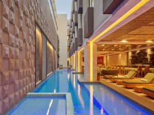 Una moderna piscina de hotel se extiende junto a un elegante salón con tumbonas verdes, mesas de madera y un bar, perfecto para eventos o reuniones, iluminado con una cálida luz amarilla y rodeado de detalles arquitectónicos geométricos.