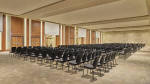 Una amplia sala de conferencias vacía en uno de los mejores complejos turísticos de México, con filas de sillas negras, suelo alfombrado de color beige, paredes revestidas de madera, pantallas de proyección y una pared decorativa de piedra: ideal para reuniones y eventos de conferencias.