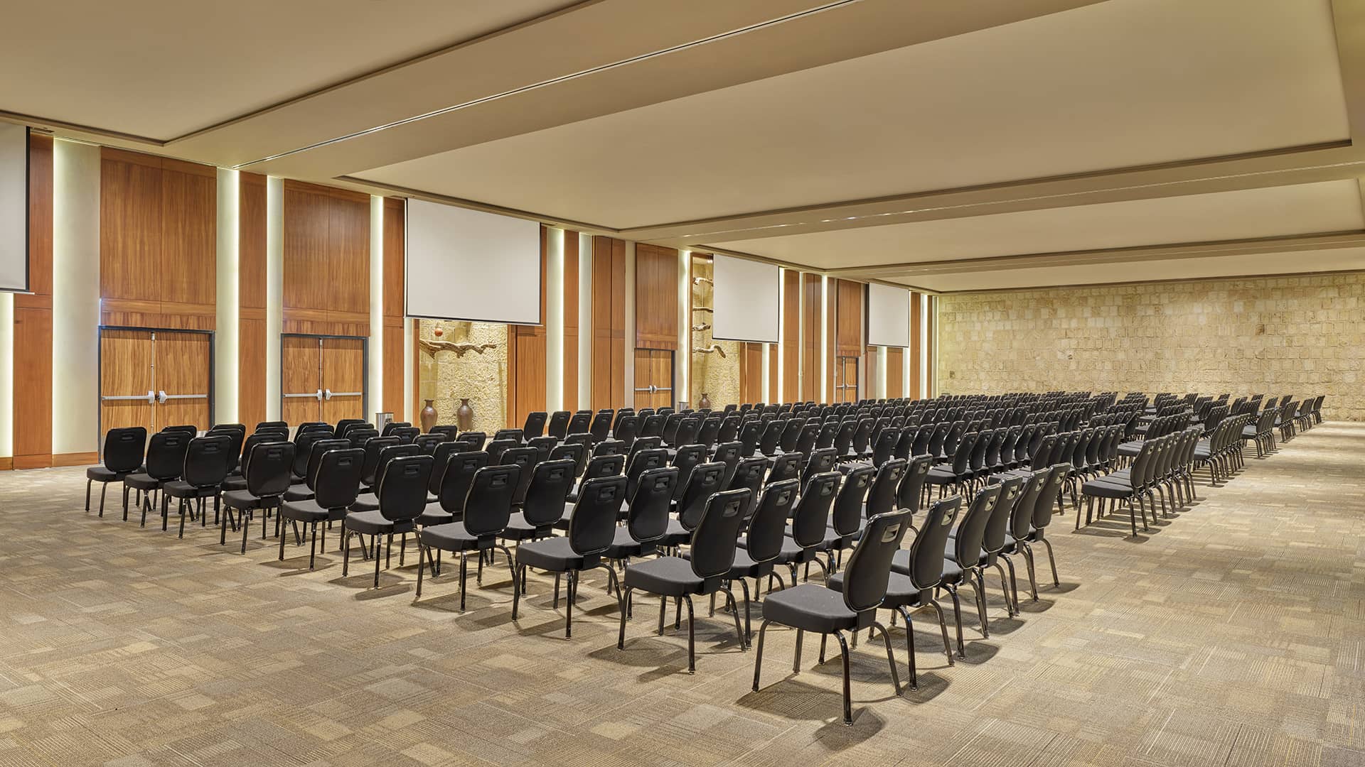 A large, empty conference room at one of Mexico’s top resorts, featuring rows of black chairs, beige carpeted floor, wooden panel walls, projection screens, and a stone accent wall—ideal for meetings and conference events.