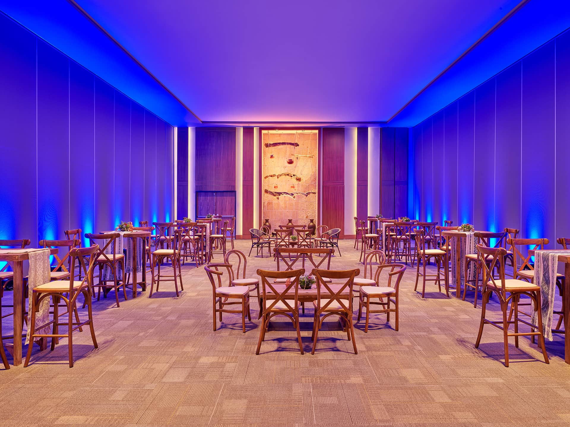 A spacious, modern event hall ideal for conference events and meetings, with small round tables and wooden chairs. The room features blue accent lighting and a large decorative wall piece at the far end, perfect for Mexico hotels or resorts.