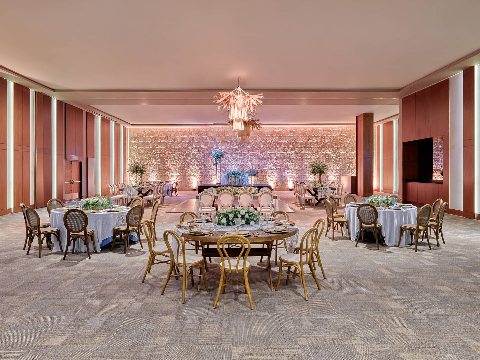 Spacious, elegant banquet hall at one of Mexico’s premier resorts with round tables and chairs, neutral tones, soft lighting, a stone accent wall, large floral centerpieces, and a modern chandelier—perfect for meetings or conference events.