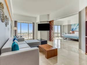 Amplia y moderna suite de hotel en uno de los mejores resorts de México, que cuenta con un sofá modular gris, cojines decorativos azules, mesas de centro de madera, un televisor grande, ventanales del suelo al techo, balcón privado y un dormitorio contiguo, perfecto para reuniones o eventos de conferencias.