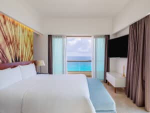 Una moderna habitación de hotel en uno de los mejores hoteles de México, con una cama grande, un banco azul, un televisor de pantalla plana y un ventanal con vistas al mar. Los tonos neutros y la iluminación tenue crean un ambiente tranquilo, ideal para reuniones o conferencias.