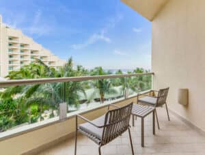 Un balcón de hotel con dos sillas y una mesita ofrece vistas a palmeras, un edificio del complejo turístico y el océano bajo un cielo azul despejado; perfecto para relajarse después de eventos de conferencias en hoteles de México.