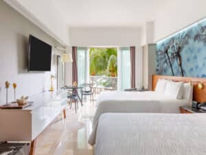Habitación de hotel moderna con dos camas dobles, televisor de pantalla plana, escritorio para reuniones y balcón con sillas con vistas a exuberantes palmeras. El espacio es luminoso gracias a la luz natural y cuenta con una decoración contemporánea, ideal para eventos y conferencias en resorts de México.