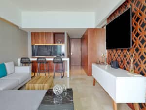 Sala de estar moderna con un sofá seccional gris claro, mesas de centro de madera, un televisor de pantalla plana en una pared decorativa estampada y un mueble para equipos multimedia blanco; perfecta para reuniones informales o para relajarse después de eventos de conferencias en los mejores hoteles y resorts de México.