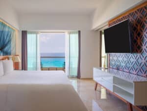Habitación de hotel moderna con una cama grande, televisor de pantalla plana y puertas corredizas de cristal que dan a un balcón con vista al mar. Ideal para conferencias o reuniones, este resort en México cuenta con muebles elegantes y pisos de mármol.