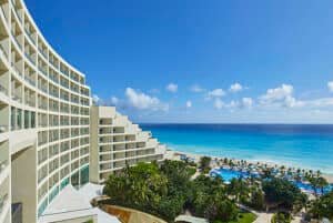 Un moderno hotel resort frente al mar en México, con balcones que ofrecen vistas a un océano turquesa, una playa de arena, palmeras y una gran piscina al aire libre, perfecto para reuniones o eventos de conferencias bajo un cielo azul brillante con nubes dispersas.